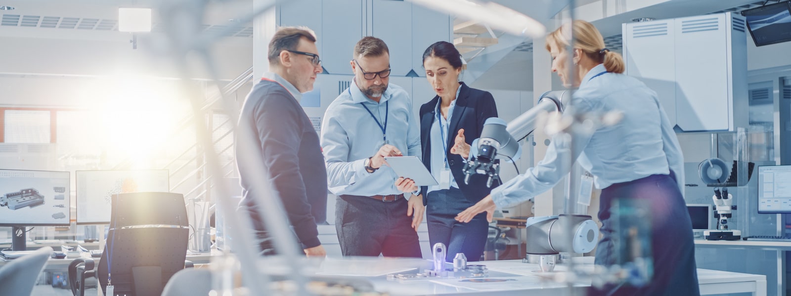 Diverse Team of Industrial Robotics Engineers Gathered Around Table With Robot Arm, They Use Tablet Computer to Manipulate and Program it to Pick Up and Move Metal Component. Bright Facility